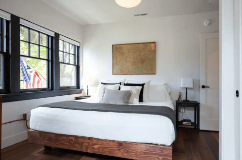 A cozy bedroom featuring a wooden bed, large windows, and a vintage map on the wall. An American flag is visible outside.